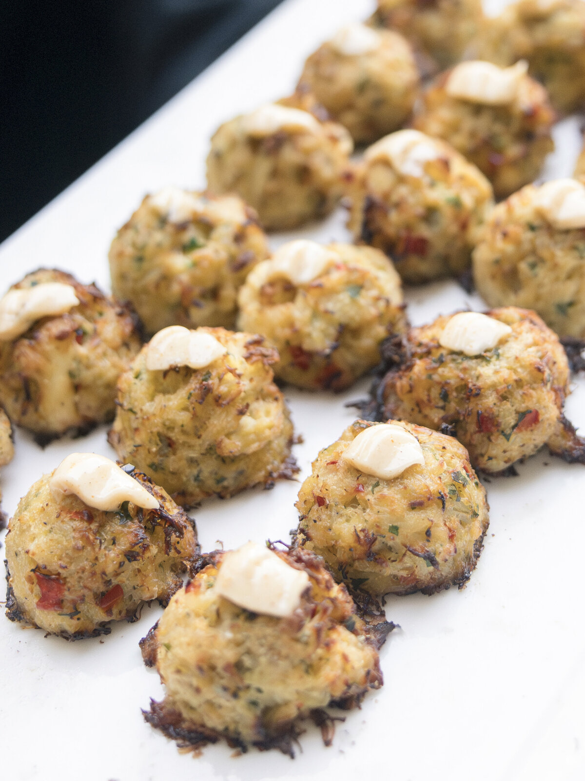 A white platter displaying several baked crab cakes topped with a dollop of creamy sauce. The crab cakes are golden brown with visible flecks of herbs and spices.