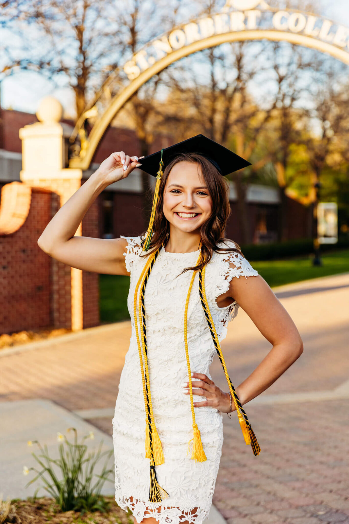 Stunning college graduate in a white dress holding her graduation cap while standing on campus during her graduation photo session