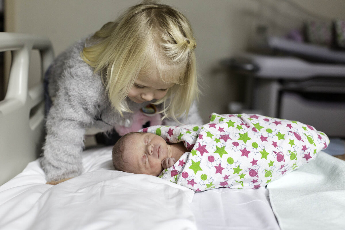 a toddler looks at her brand new baby sister in a hospital room after she is born