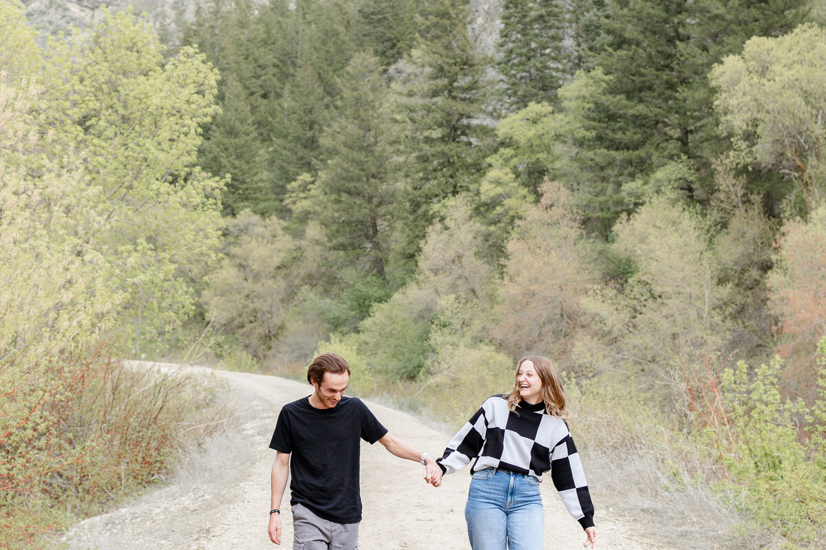 A happy couple holding hands and laughing while walking along a dirt path in Vivian Park, surrounded by lush greenery and towering trees.