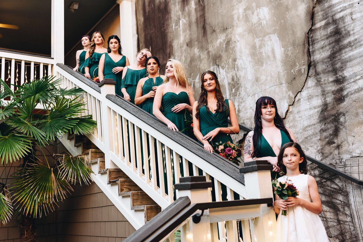 A group of bridesmaids in green dresses and a young flower girl in white stand on a staircase beside a weathered wall, holding bouquets and smiling, with greenery and string lights nearby.