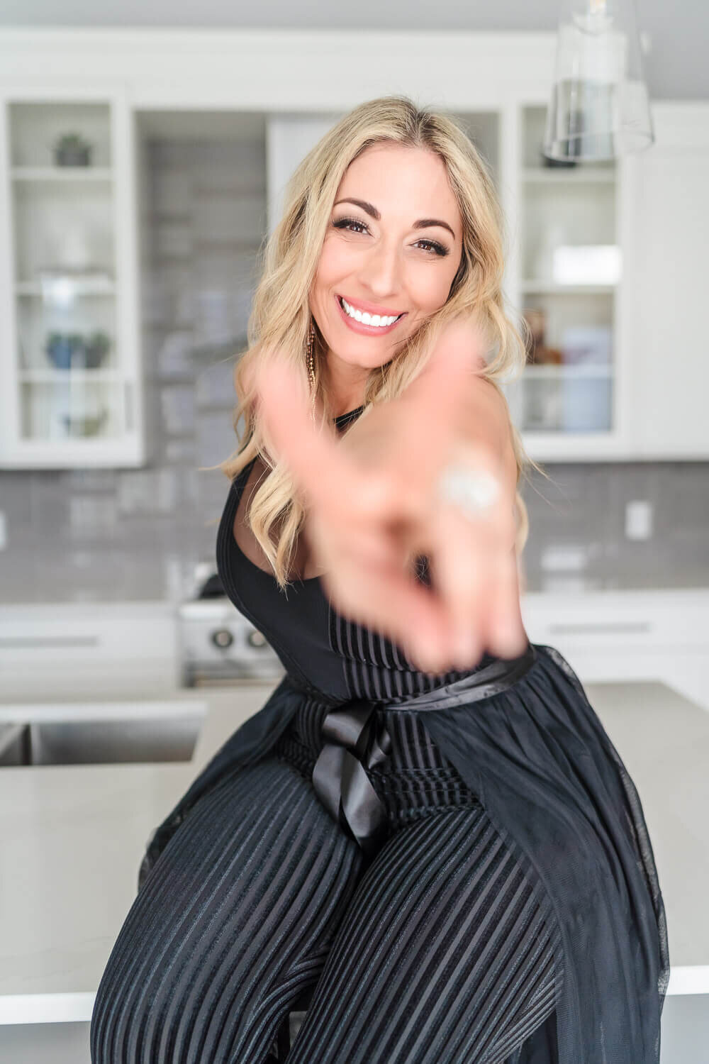 Blonde entrepreneur in black outfit smiling and giving blurred peace sign toward camera in bright modern kitchen.