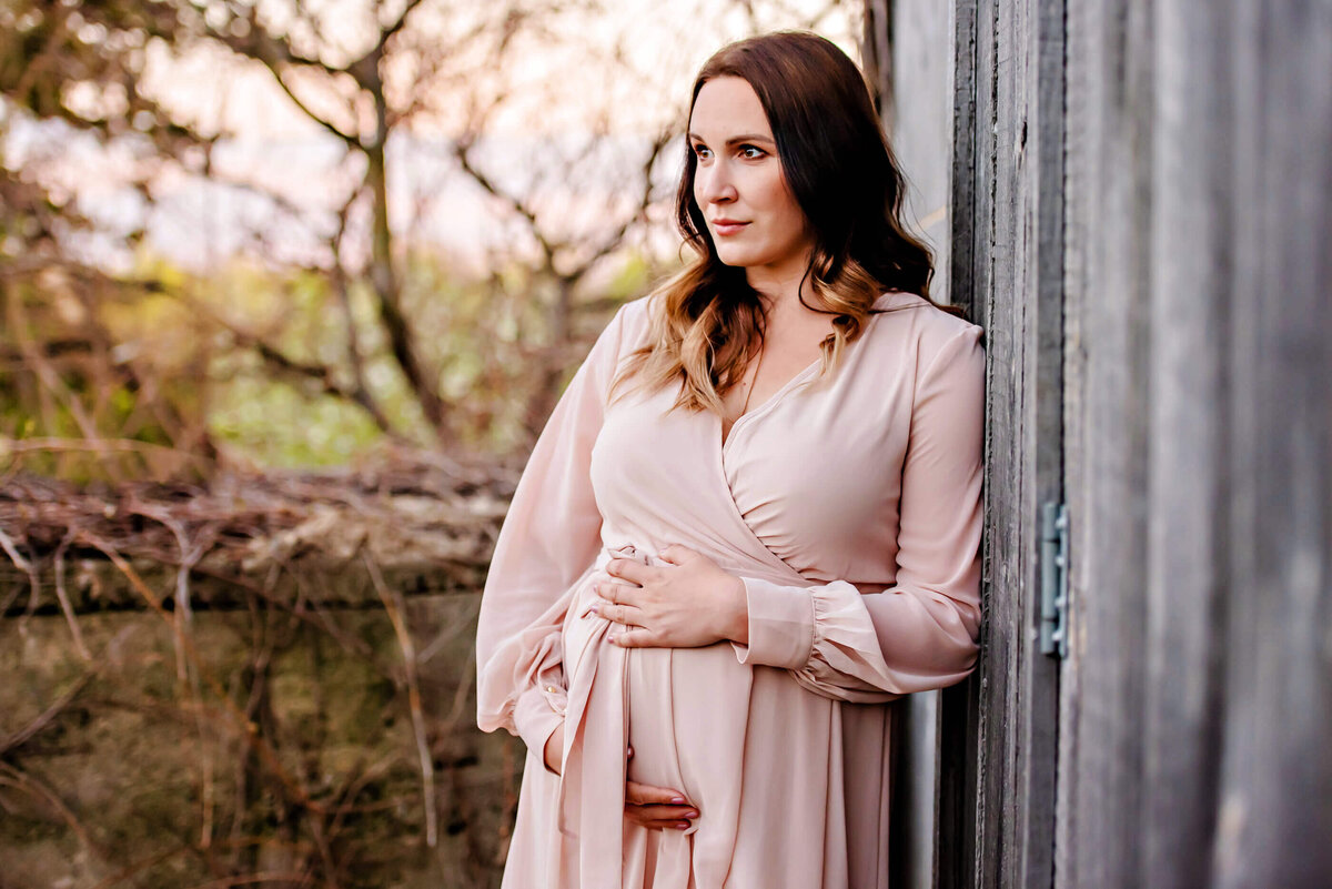 pregnant woman cradling her belly leaning against a barn for her golden-hour maternity session in Toronto, Ontario.