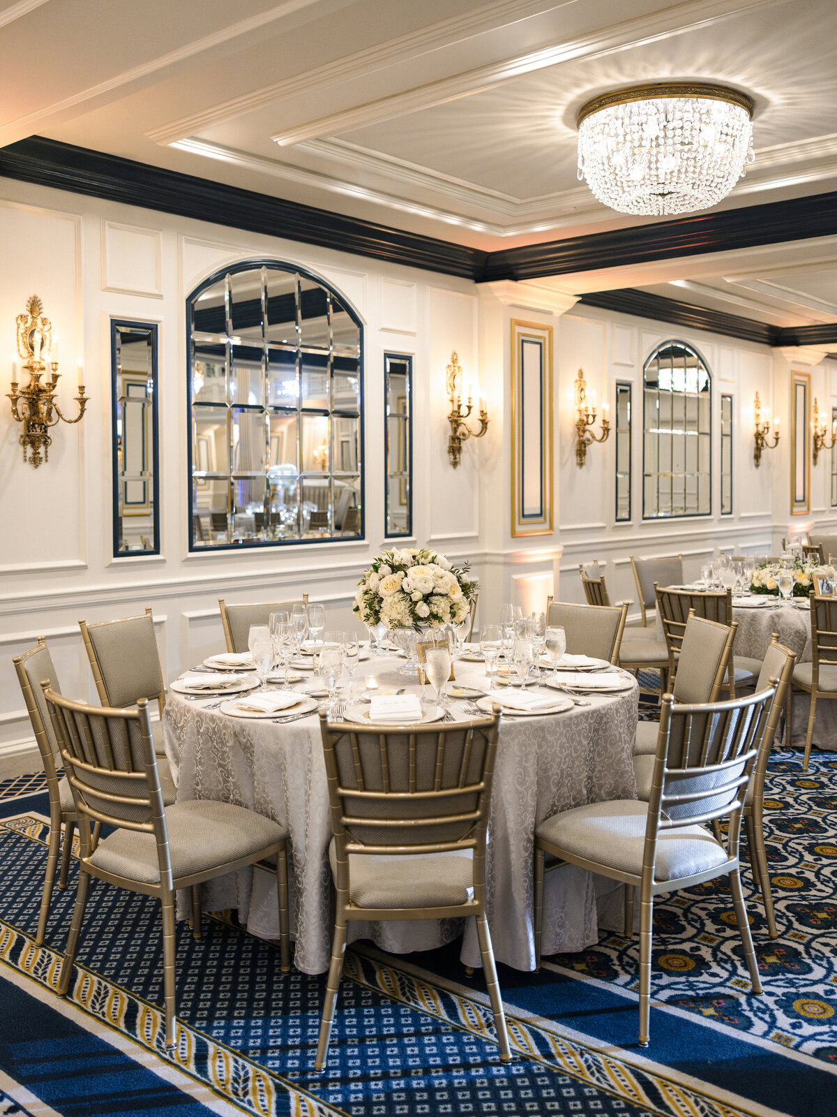 Elegant banquet hall with a round dining table set for an event. It is adorned with white linens, fine china, silverware, and a floral centerpiece. Gold chairs surround the table. Wall mirrors, sconces, and a chandelier enhance the luxurious ambiance.