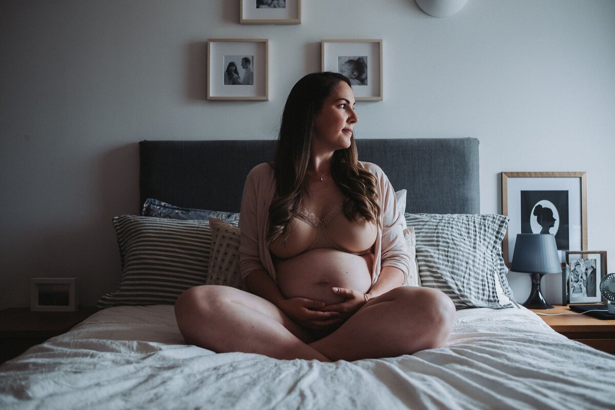 Woman wearing bra and cardigan holds her pregnant belly and looks out the window