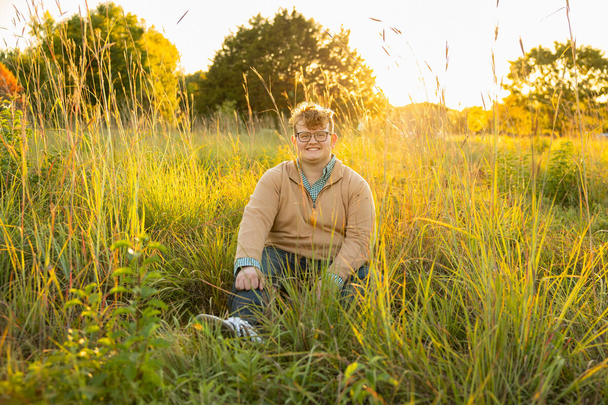 A senior guy sitting in tall grass smiling in the golden hour in Lawrence, KS
