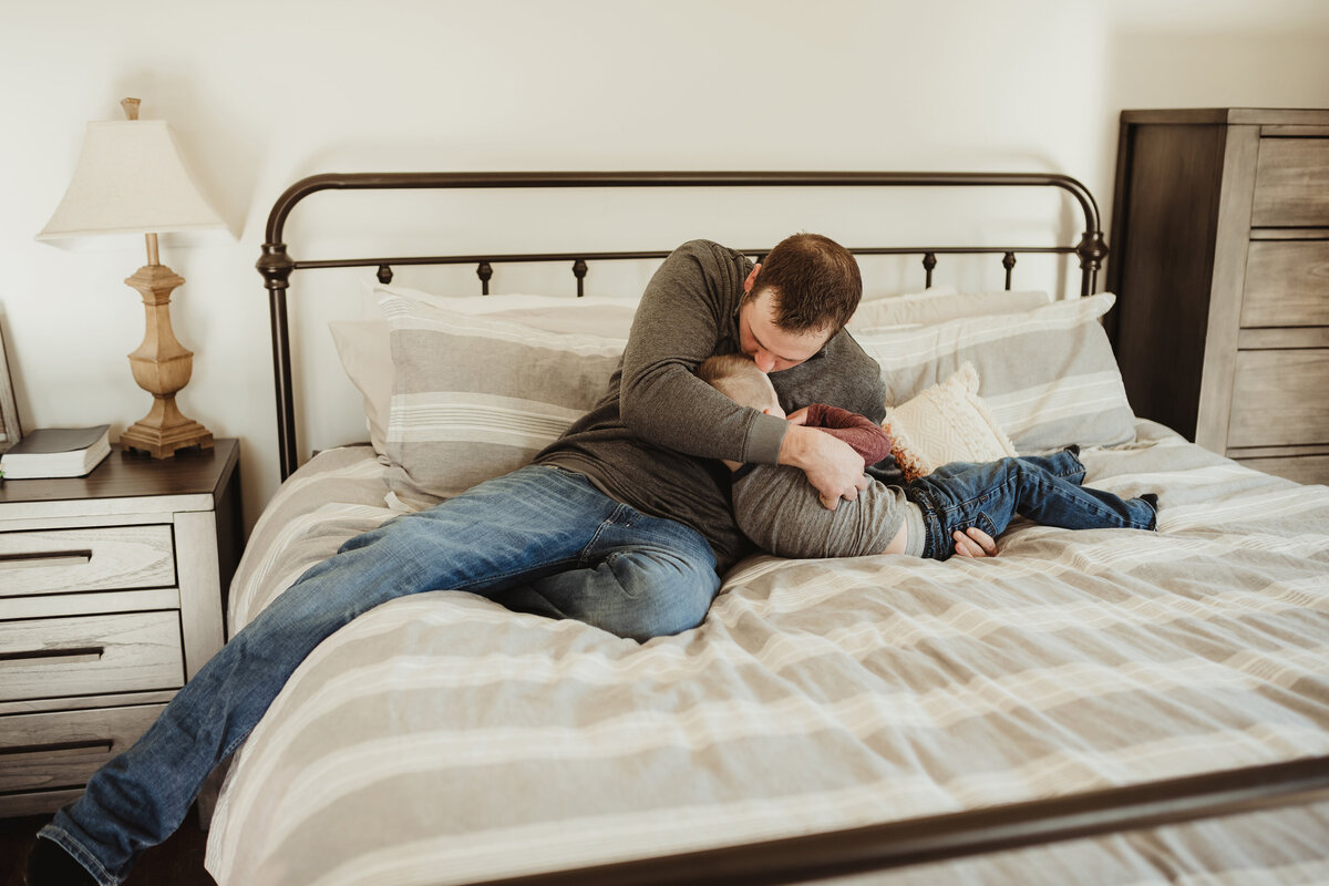 A dad is kissing and cuddling his toddler son while sitting on the bed.