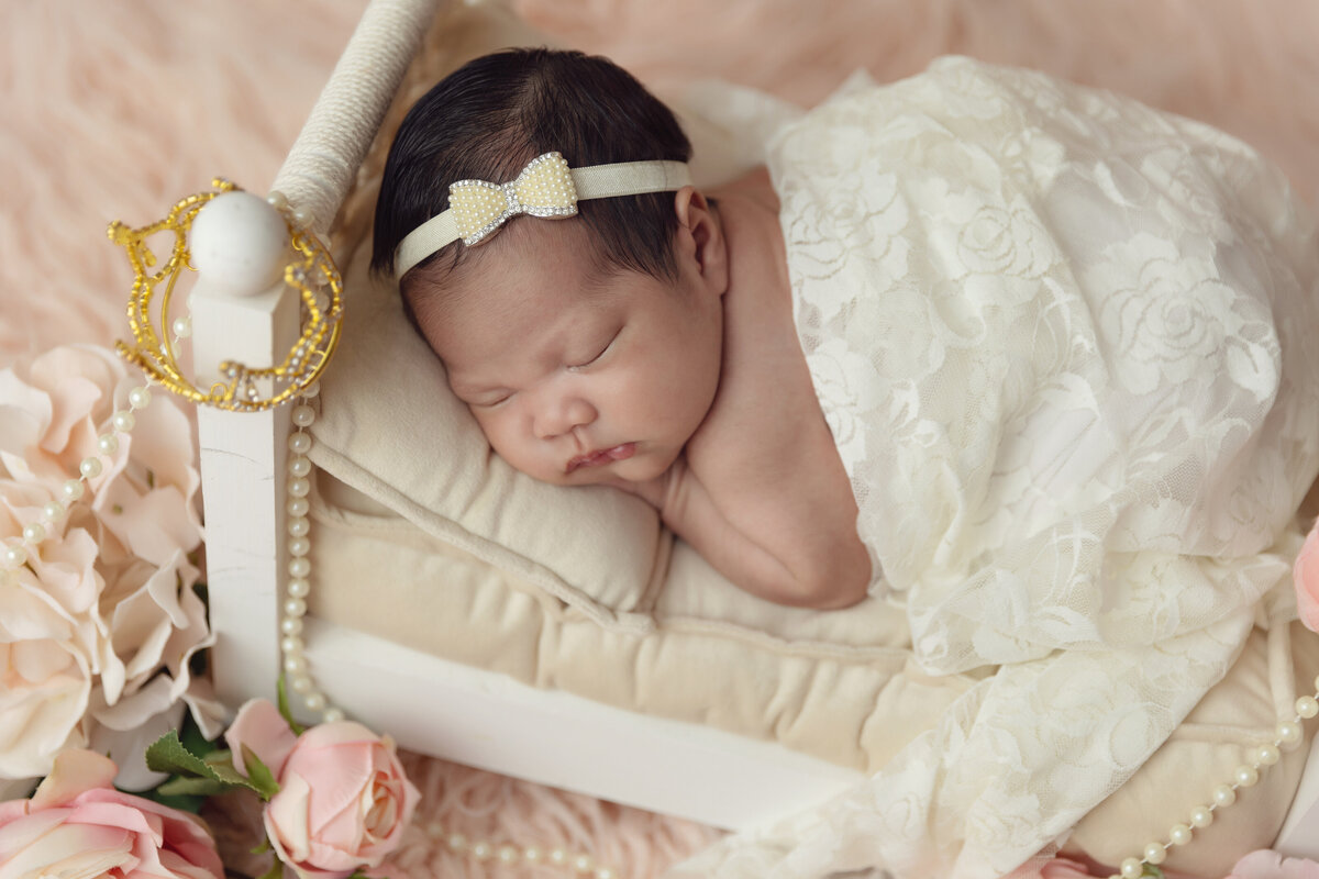 A newborn baby girl wearing a white bow sleeps under a lace blanket in a wooden bed