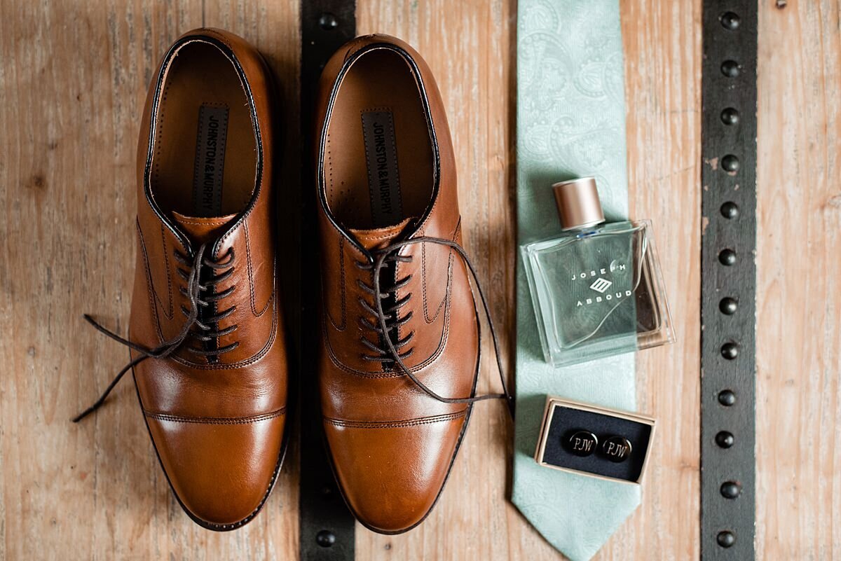 Mens brown leather shoes on a table next to a light green tie with a square bottle of cologne and a black box of shiny black cuff links.