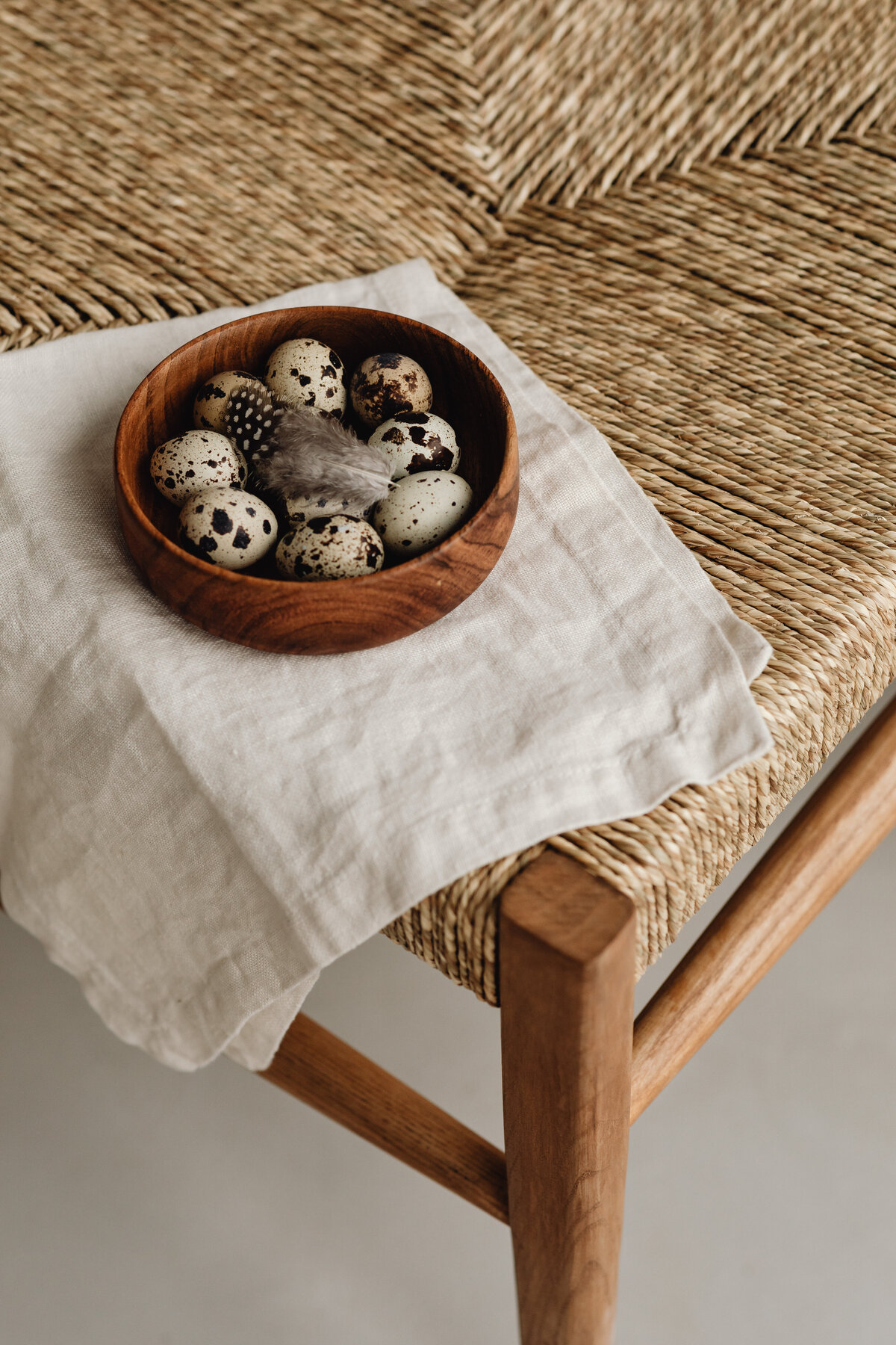 earth-tone speckled eggs and feather  in a wooden bowel on a wicker chair and linen cloth