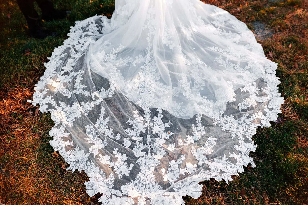 A close-up of a wedding dress train spread across grass, featuring intricate white floral lace embroidery and delicate sheer fabric, beautifully captured by an experienced NJ wedding photographer.