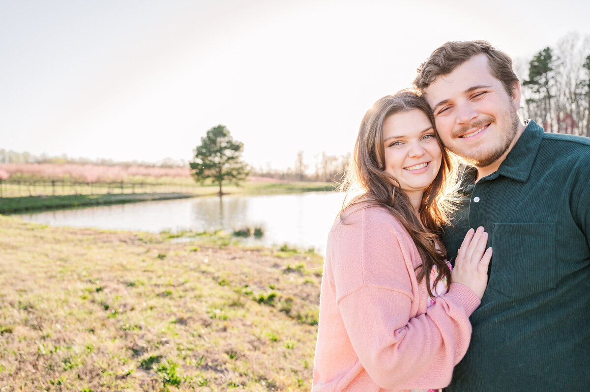 A stunning brunette engaged couple hugging at sunset next to a pond and bright cherry blossoms enjoying their destination wedding photography session with JoLynn Photography