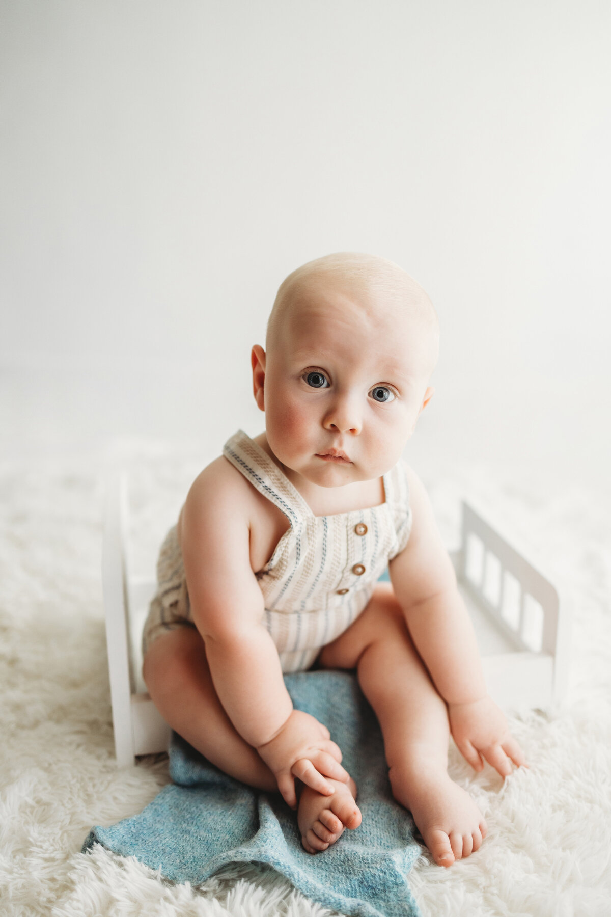 Infant sitter baby boy sitting in small wooden bed in linen overalls for his milestone photo session in Denver