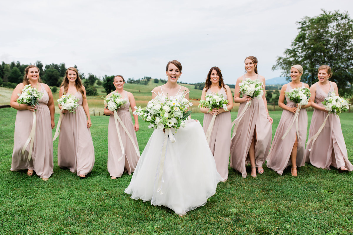 A bride walks towards the camera in a white wedding gown holding a bouquet of white flowers while seven ladies in pale pink dresses walk behind her and smile at the camera also holding bouquets
