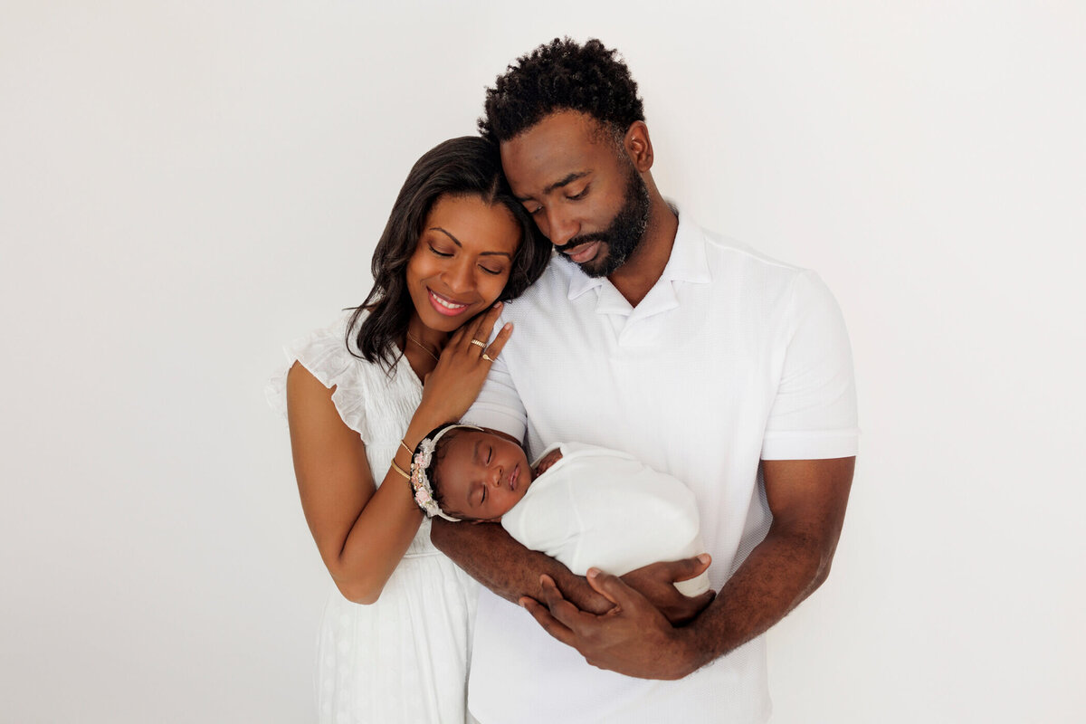 A smiling couple in white clothes lovingly looks at their newborn baby, swaddled in white, as the father gently holds the baby in his arms.