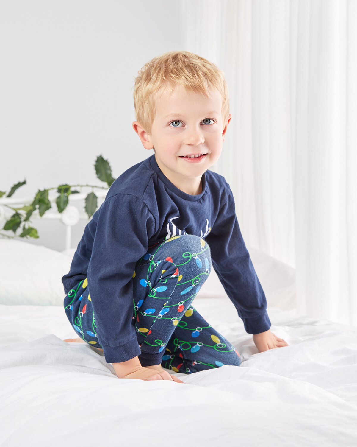 Image of a boy in his pyjamas on a white bed with holly Christmas backdrop in the studio. Taken by Norwich portrait photographer Claire Howes. Christmas Mini Sessions Norwich.