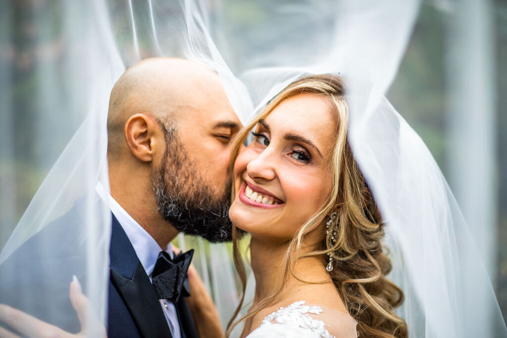 An image of a groom kissing a bride while her white veil flows around her