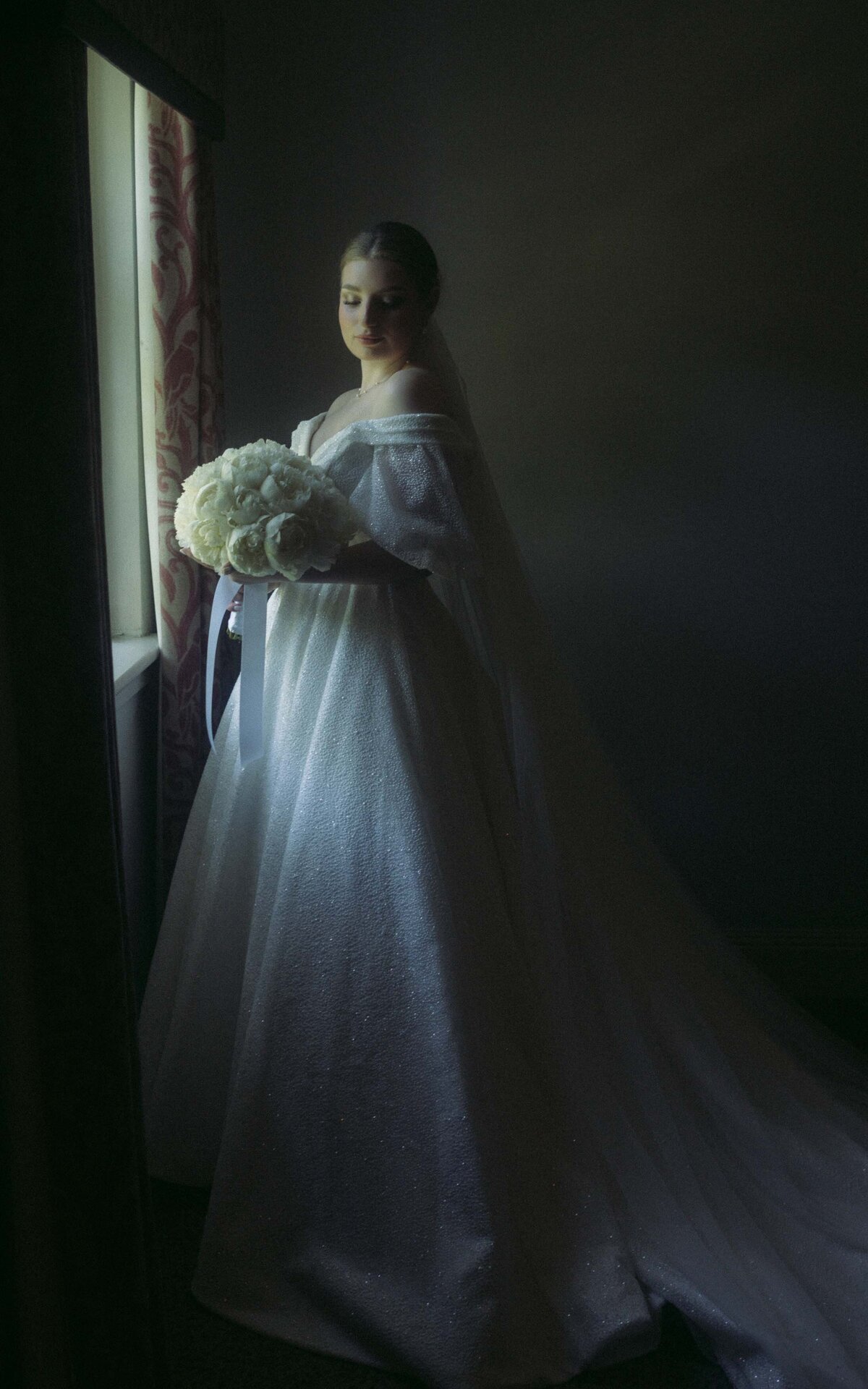 Capture the serene beauty of the bride in soft light, standing next to the window on her wedding morning, radiating elegance and anticipation for her special day