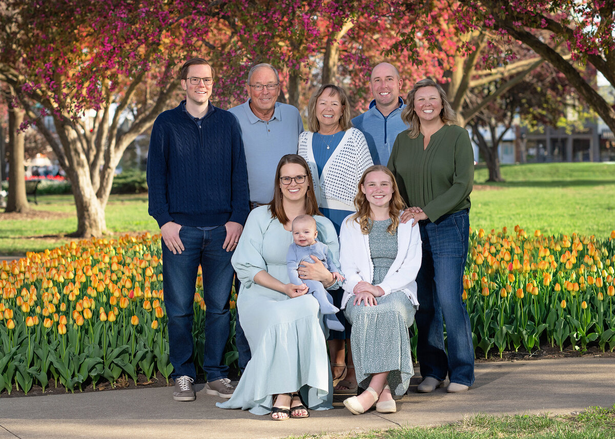 A multi-generational family, including a baby, is posing together for a portrait outdoors with a backdrop of blooming trees and yellow tulips.