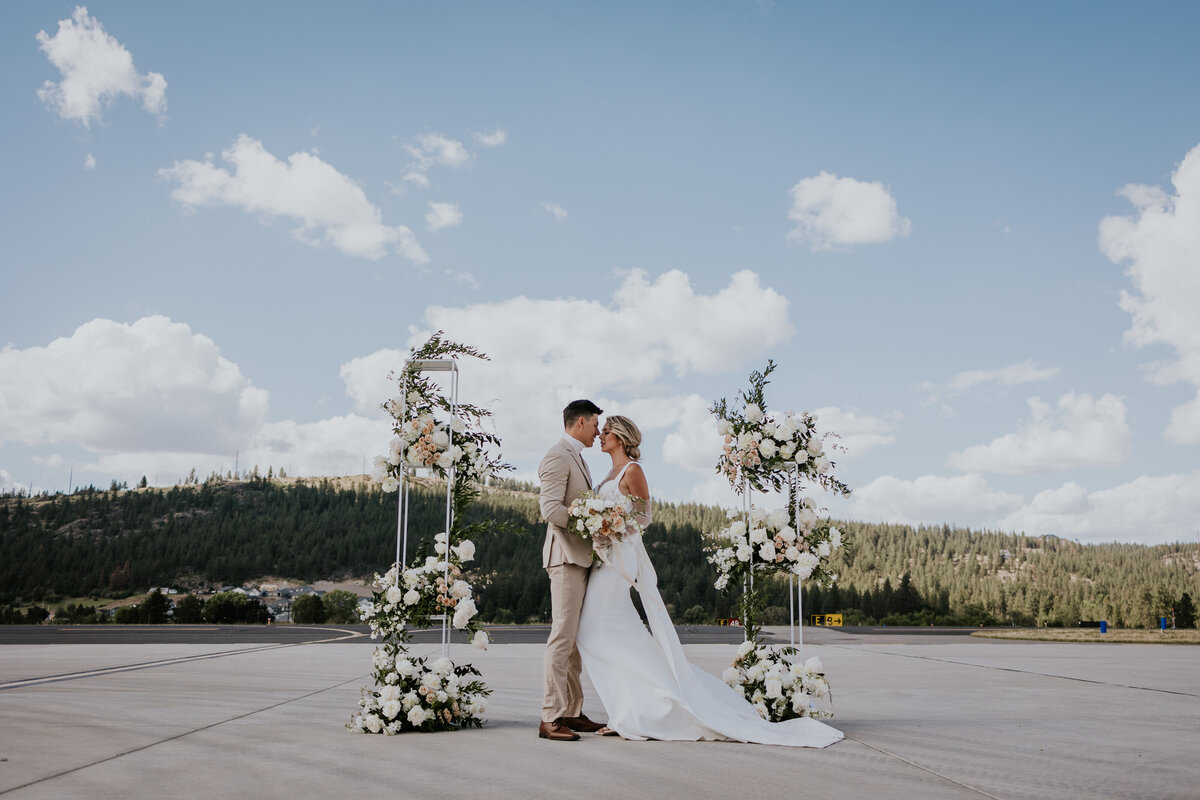 Bride and groom lean in for kiss on plane tarmac with flowered arbor behind them.