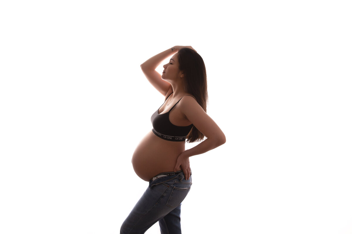 Pregnant woman wearing jeans and a black bralette running her right hand through her hair with her left hand on the small of her back.