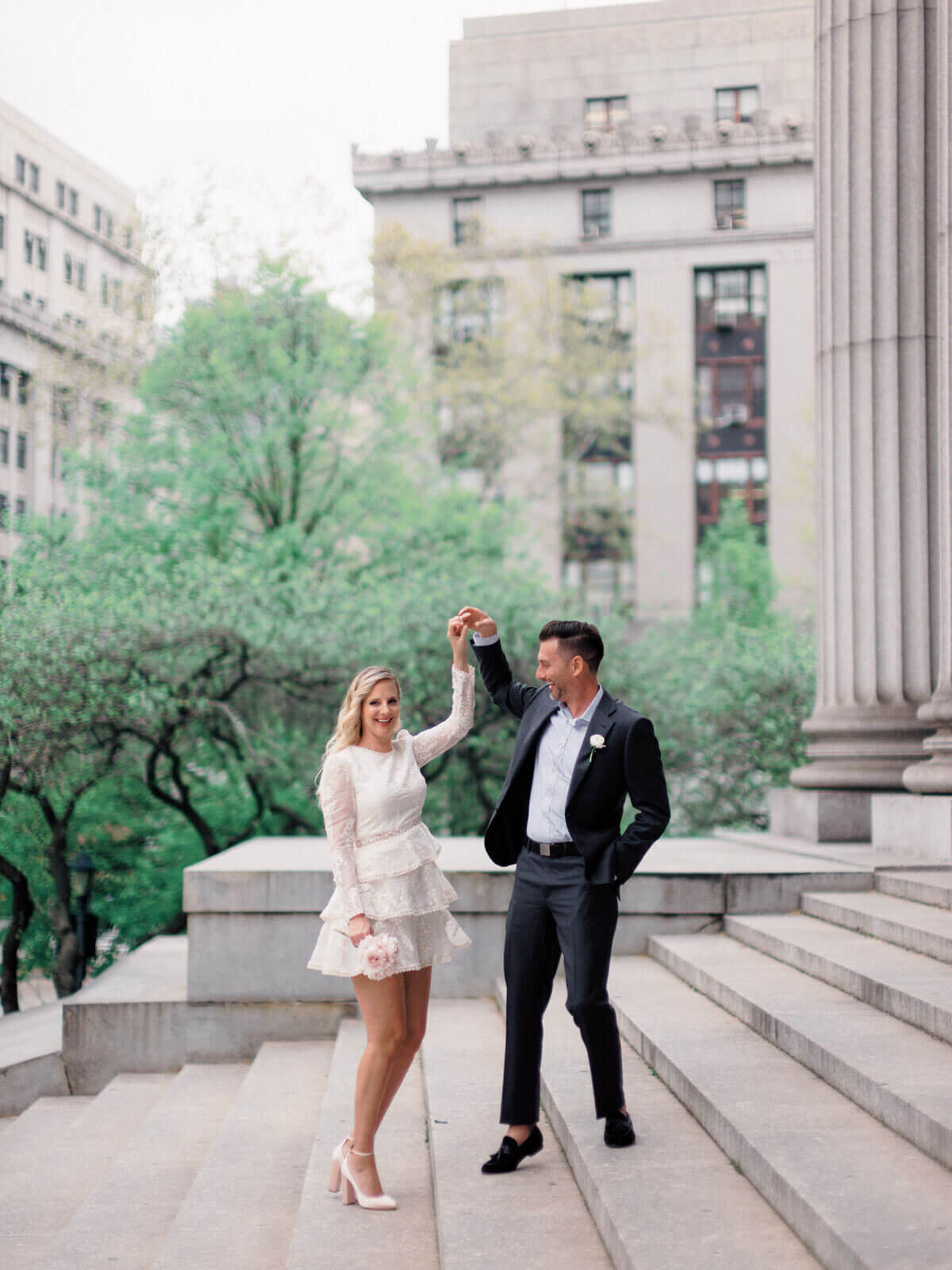 The eloping bride and groom are happily holding hands up in the air on the grand staircase of the NY City Hall. Image by Jenny Fu Studio