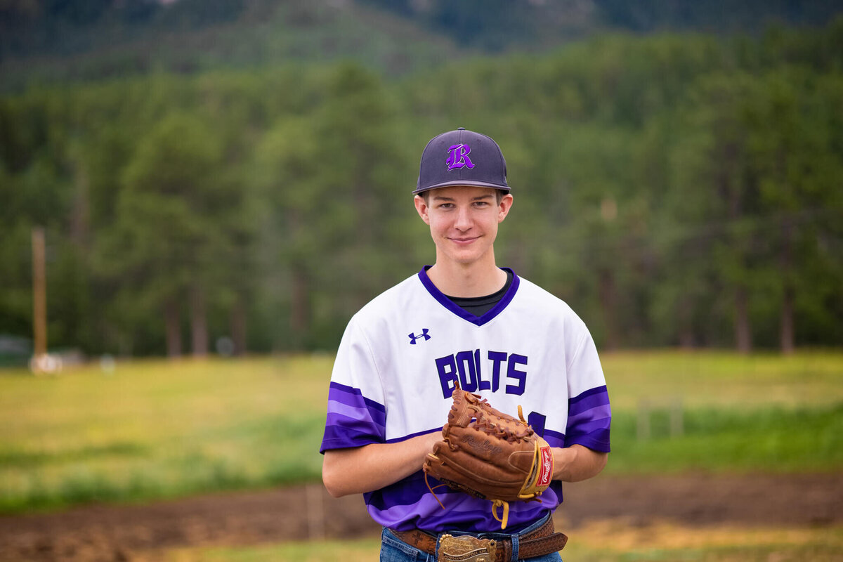 high school senior boy wearing his baseball uniform with his glove standing in a grassy field