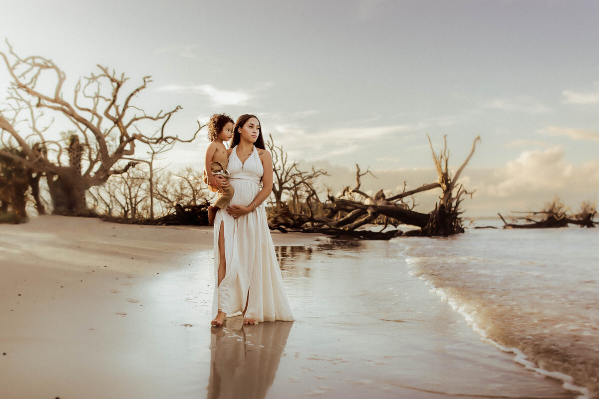 Woman in a white dress stands on a beach with a child, surrounded by driftwood and calm waves under a clear sky.
