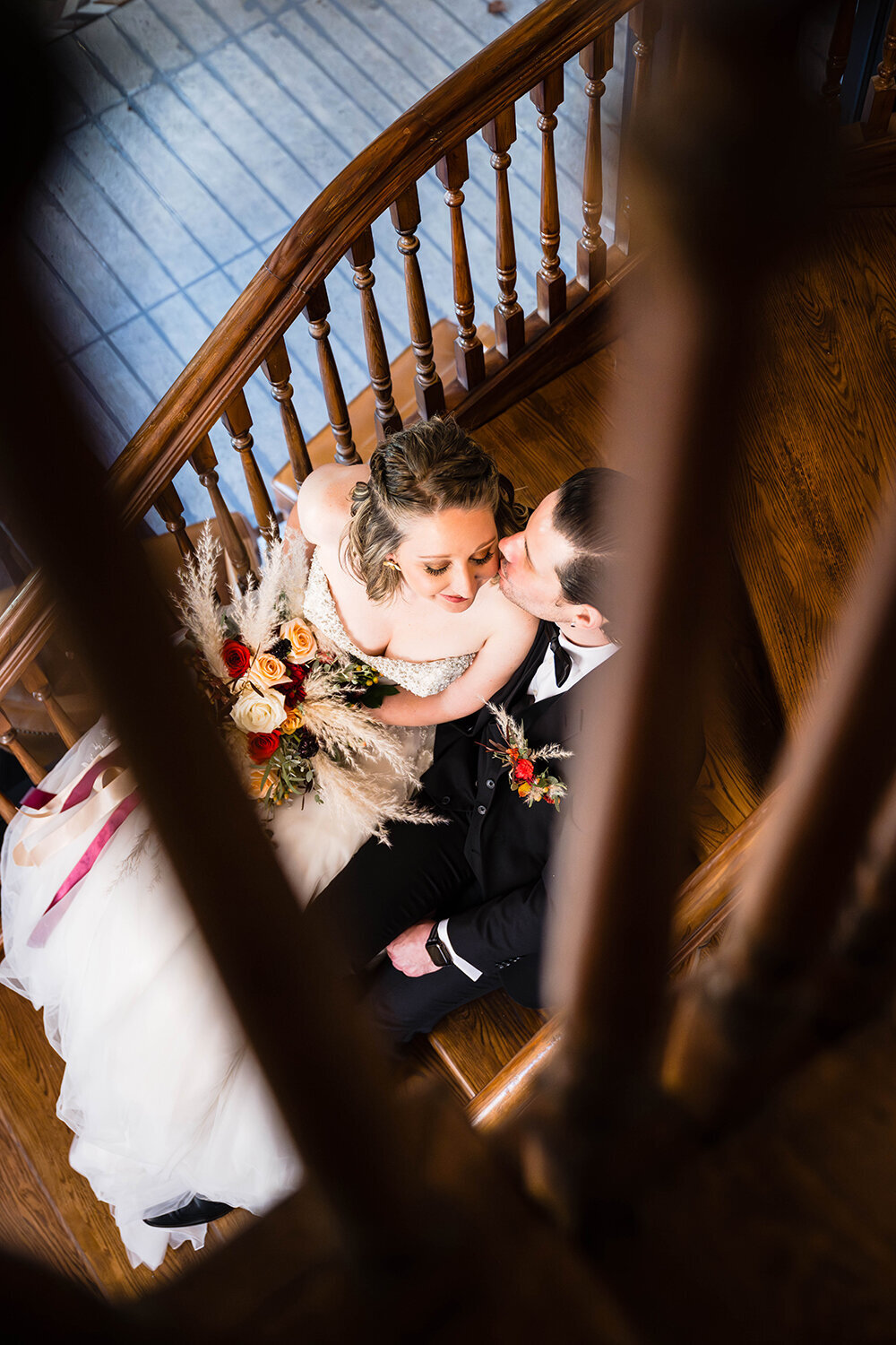 A couple on their elopement day sit side-by-side in a stairwell in a hotel located in Downtown Roanoke on their elopement day. The groom kisses the bride on the cheek while the bride smiles. A photo of them is taken through slits in the stairwell.