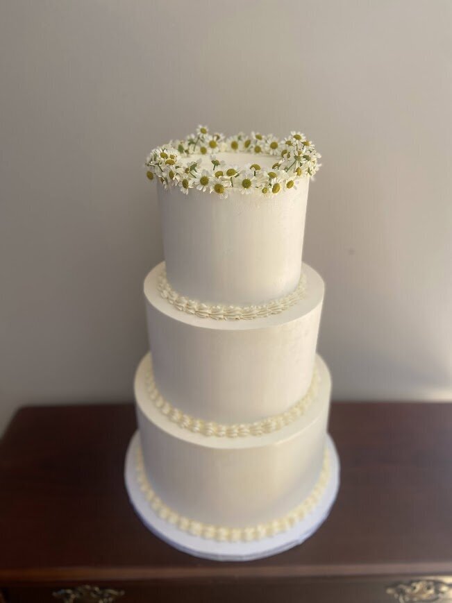 This white wedding cake is simple and clean with smooth white frosting, a piped shell border at the meeting of each tier, and chamomile flowers around the top edge of the top tier.