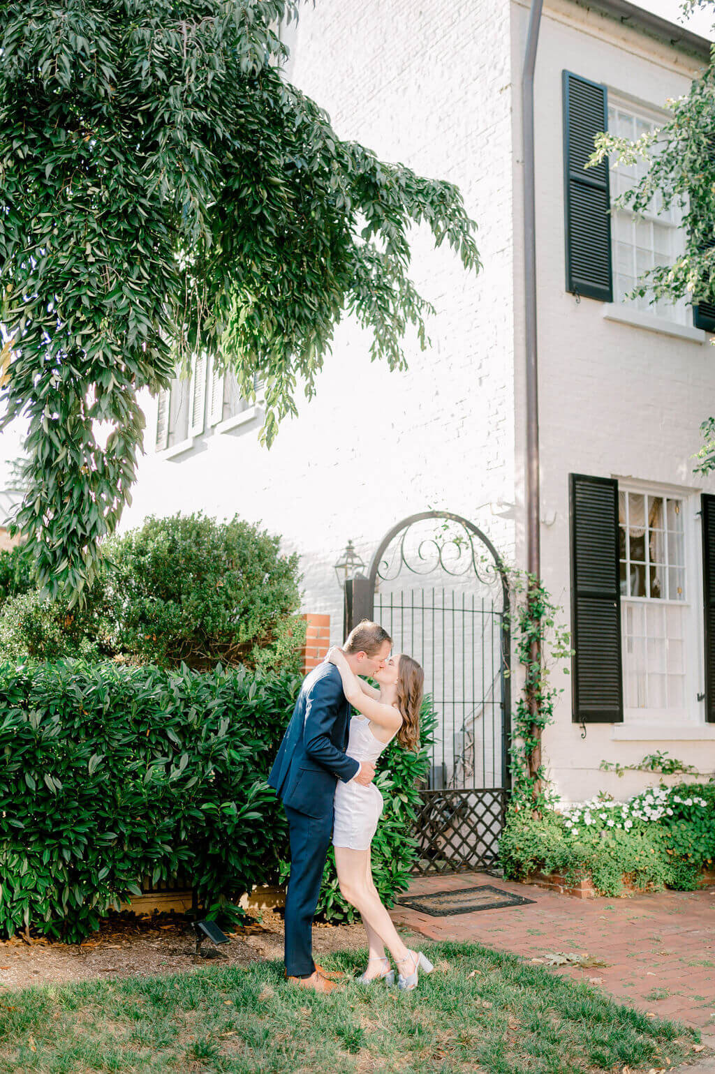 Romantic image of a couple kissing in front of a white home in old town Alexandriafor their engagement photos.