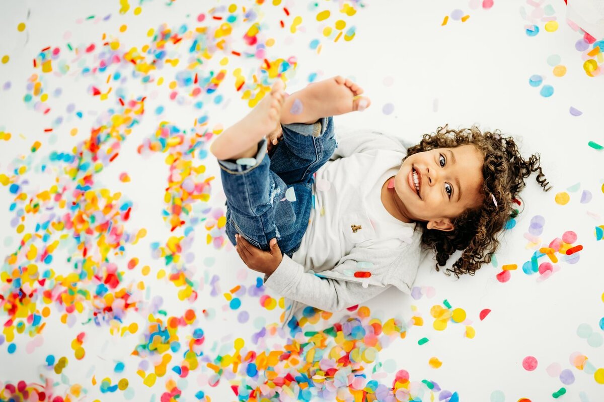 toddler boy lays in a pile of confetti looking up at the camera