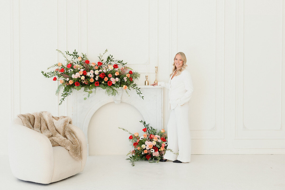 blonde woman in white suit is leaning against fireplace with pink and read floral bouquets at her feet and on the mantel. White chair nearby with cozy fur blanket