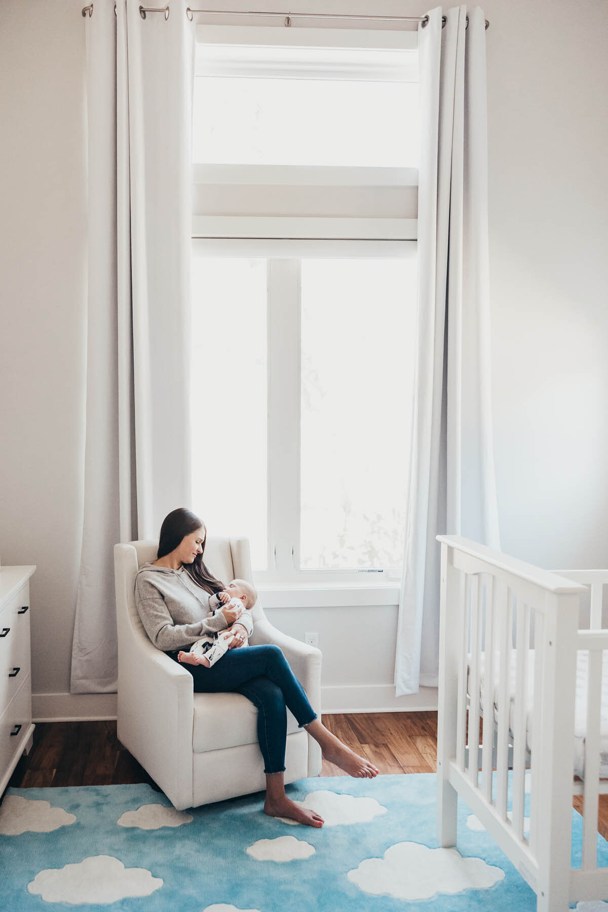 a mother holds her baby while sitting in a chair with alarge window behind her