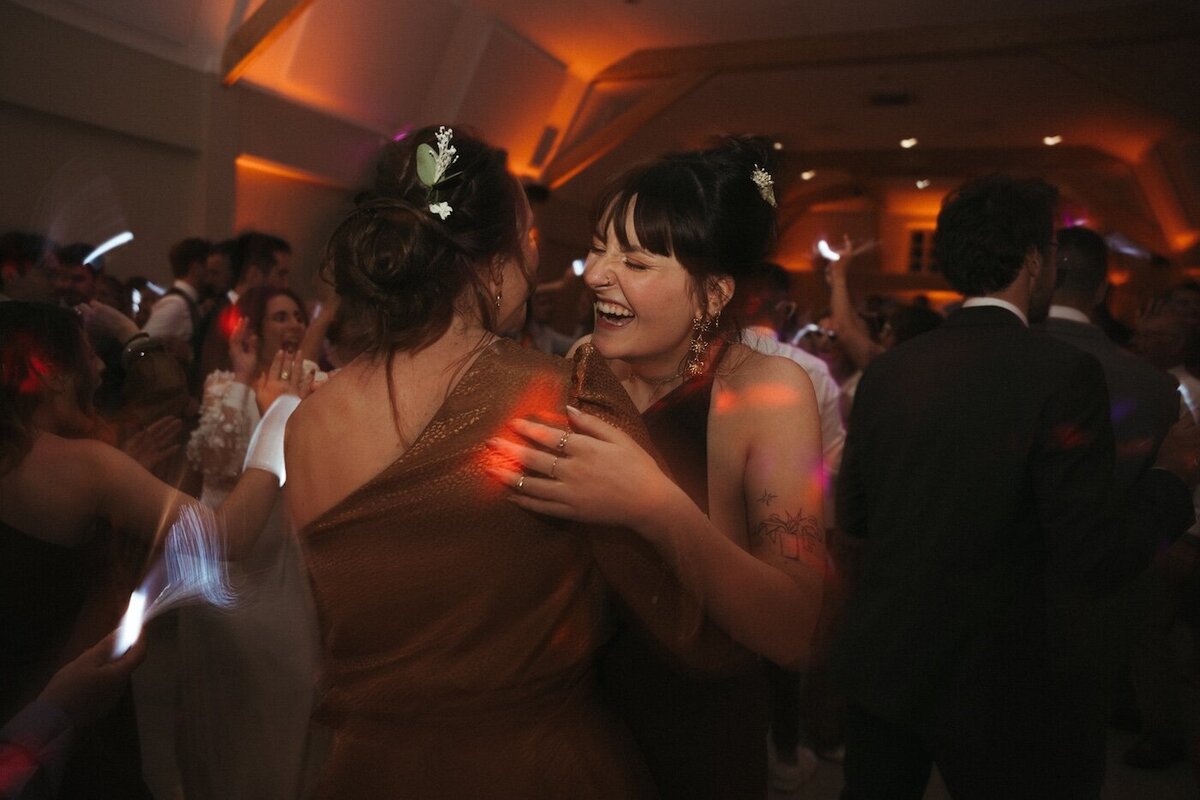 Deux invitées rient aux éclats en dansant, entourées de lumières et d'invités qui profitent de la soirée. Reportage par Kamescop, photographie et film de mariage.