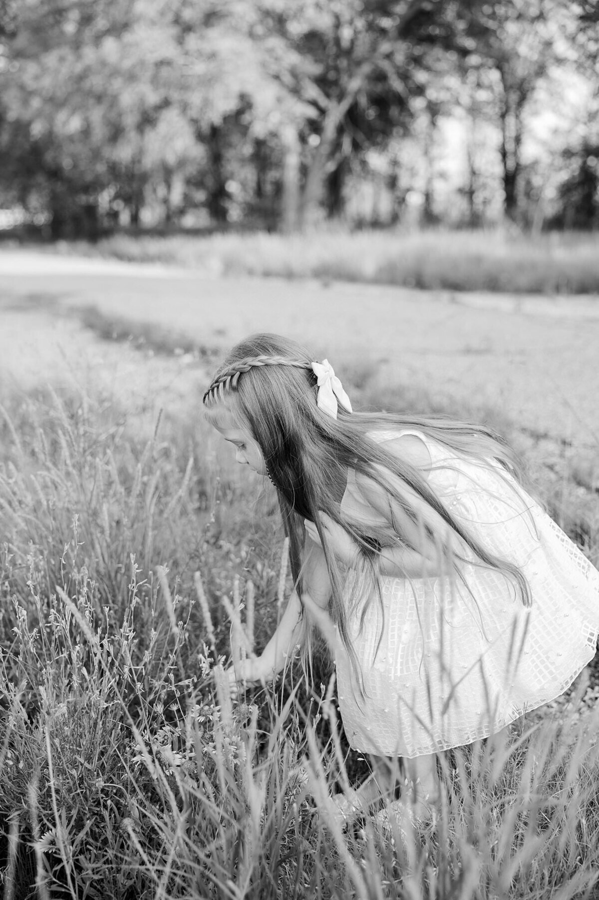 Black and white photo of a little girl bending over smelling a wild flower that is growing in a green field.