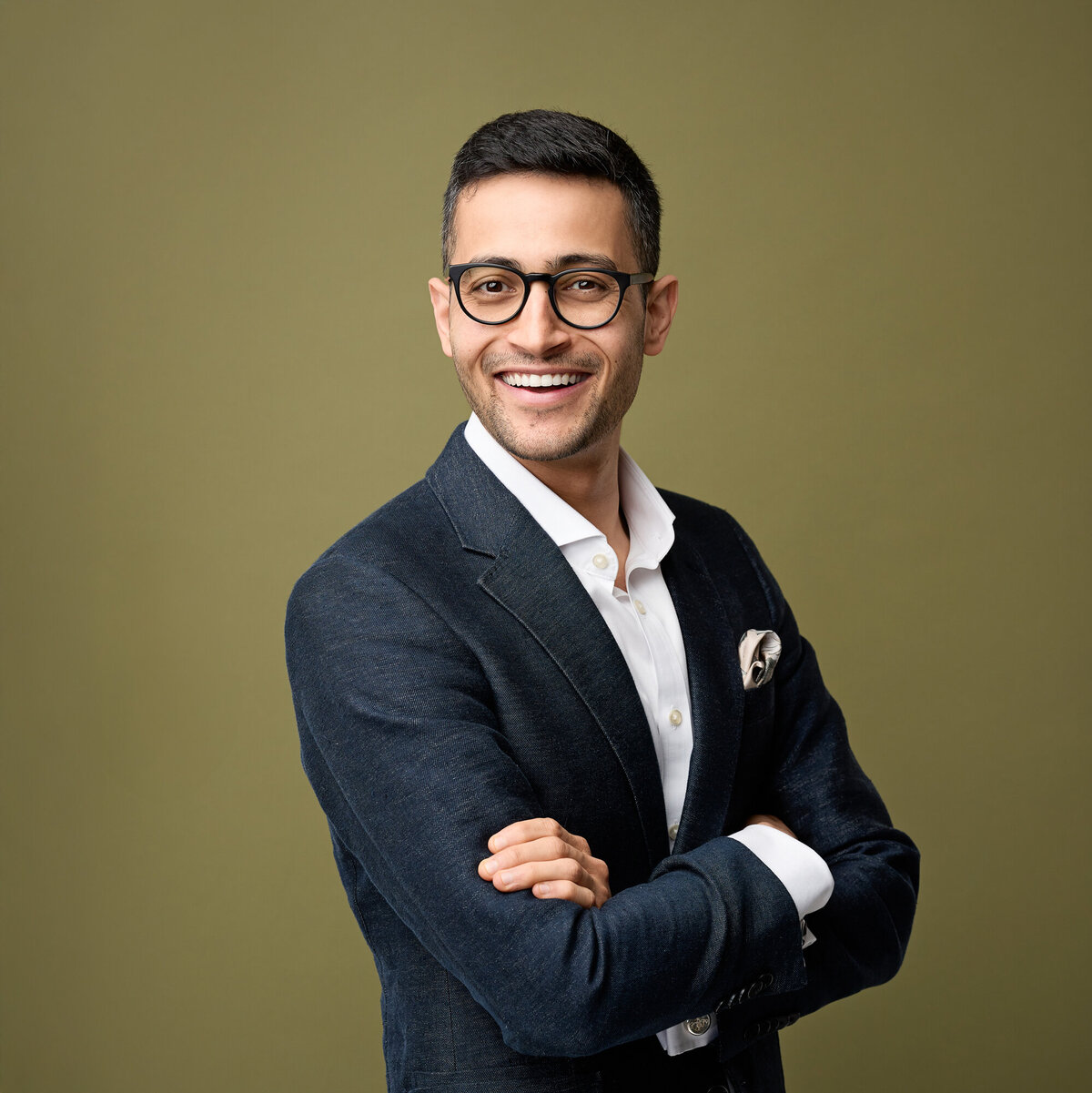Smiling man in glasses and navy jacket and white shirt. Studio photo against an olive background.