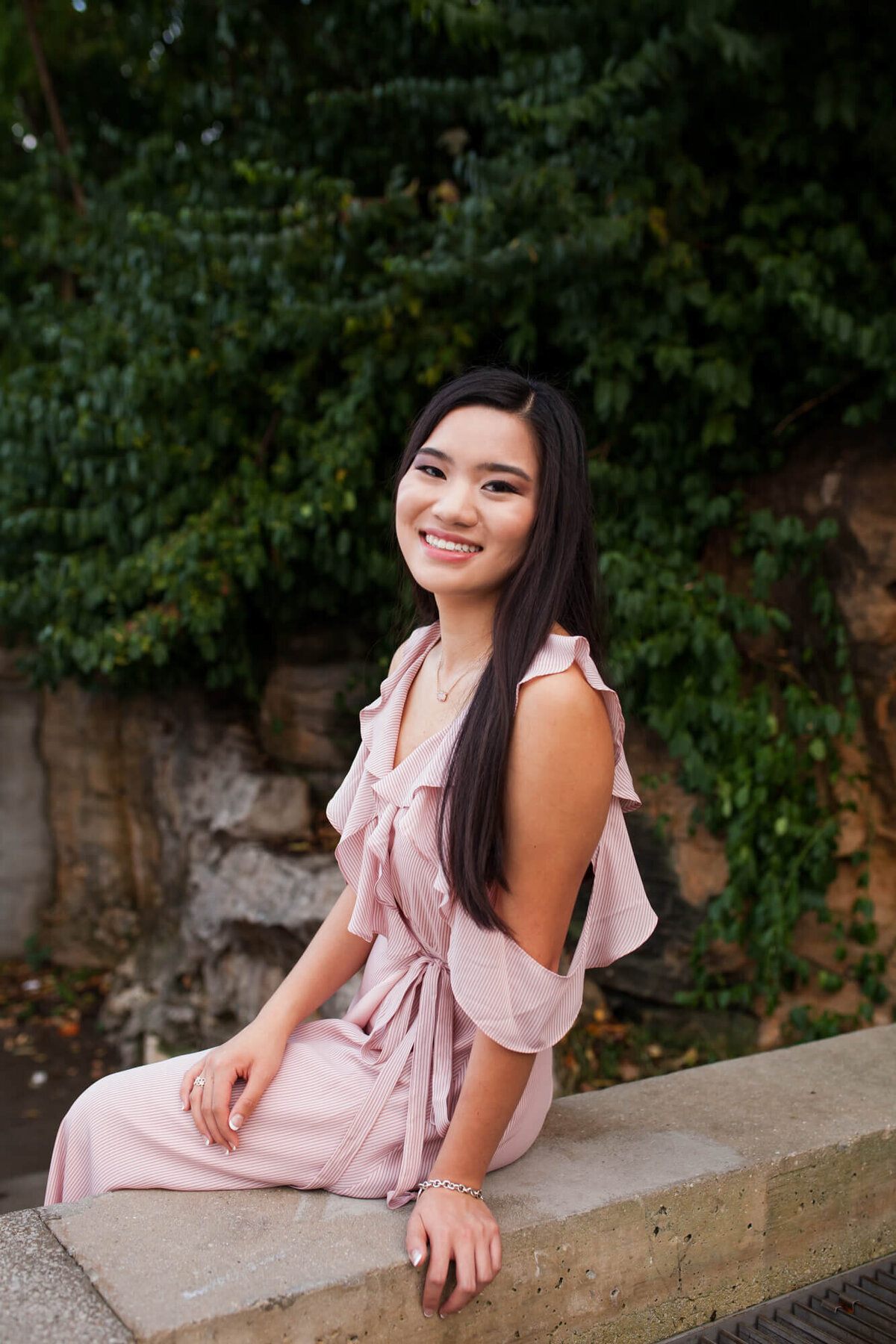A lovely dark haired teen wearing a pink ruffle top dress sits on a stone ledge for her senior portraits. Captured by Springfield, MO senior photographer Dynae Levingston.