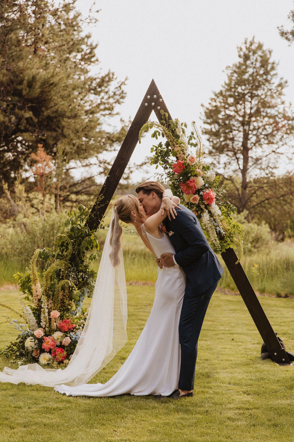 Bride and groom kissing in front of bright flowers on triangle arch on private lawn in Sunriver Oregon