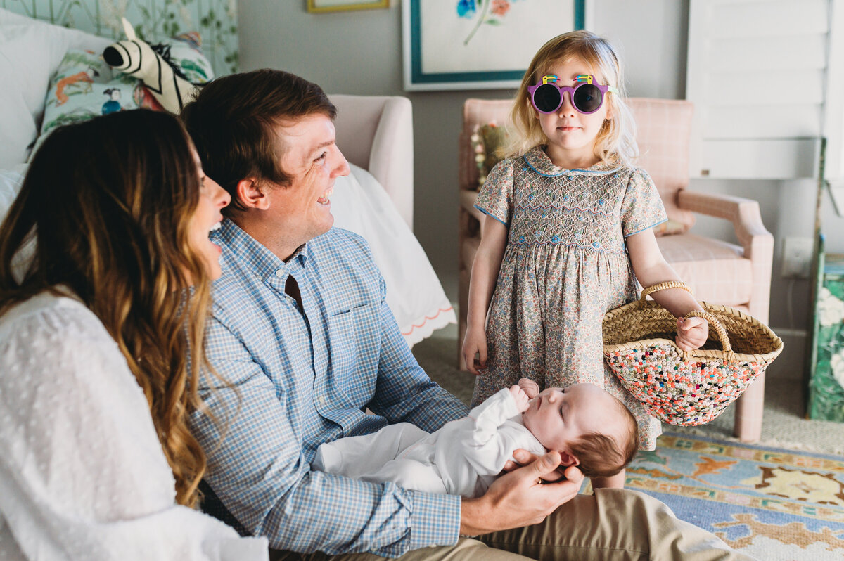 mom and dad sit on nursery floor holding new baby while big sister wears sunglasses