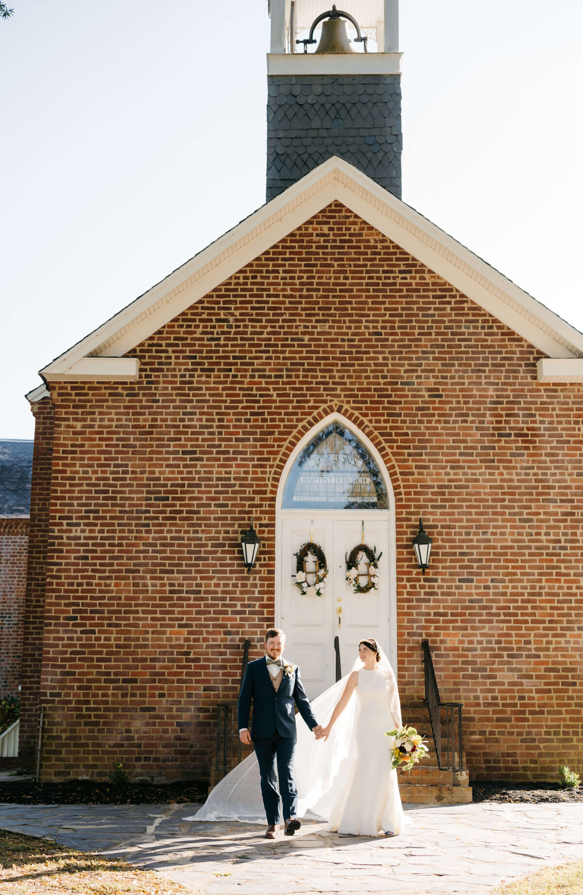richmond wedding photographer captures bride and groom holding hands as they walk in front of a brick wedding chapel with tall white doors