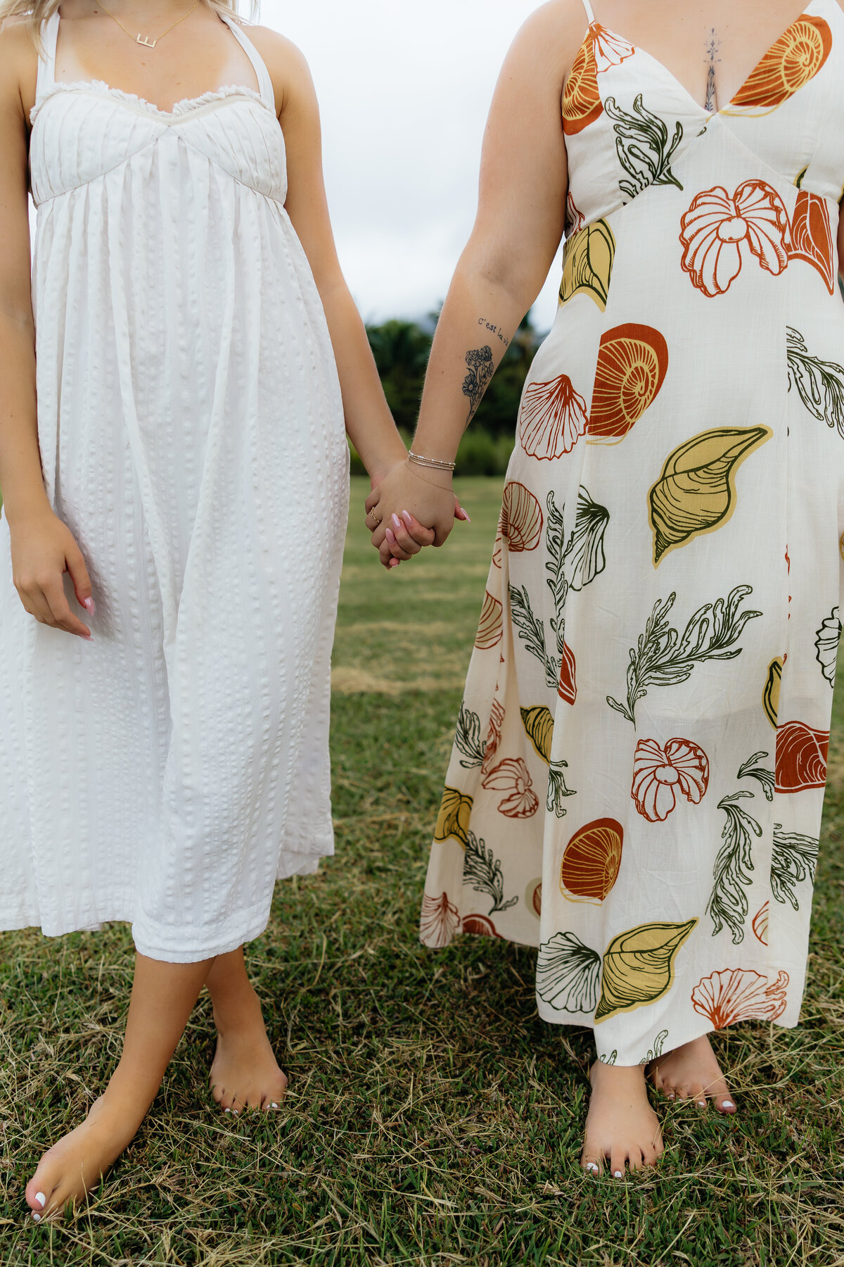Close-up of two best friends holding hands in flowy summer dresses, standing barefoot on the grass during their Oʻahu photoshoot.