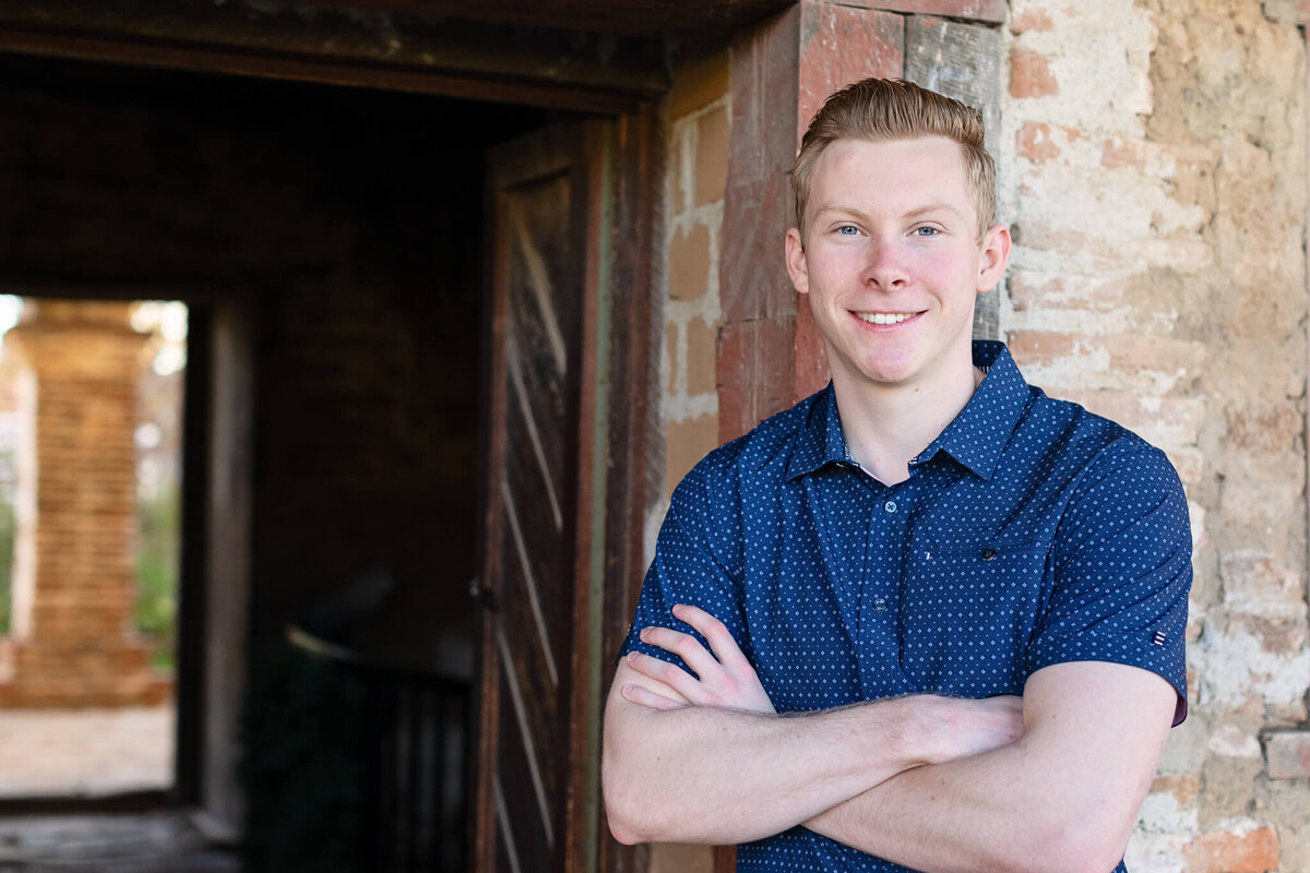 A young man with strawberry blonde hair, wearing a blue button-up shirt crossing his arms over his chest and leanign his back against a brick wall, beside a dark outdoor hallway, smiling at the camera.