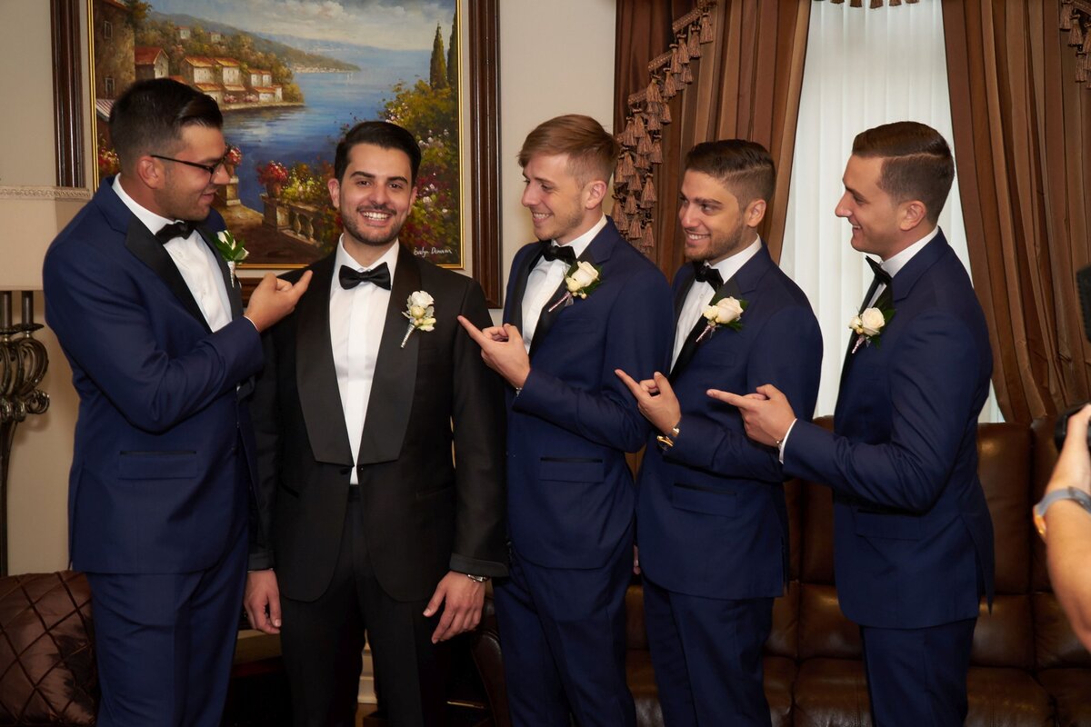 Groomsmen enthusiastically point at the smiling groom, capturing a moment of camaraderie and celebration. This image highlights the groom’s happiness and the supportive, playful interaction among his closest friends on his wedding day.