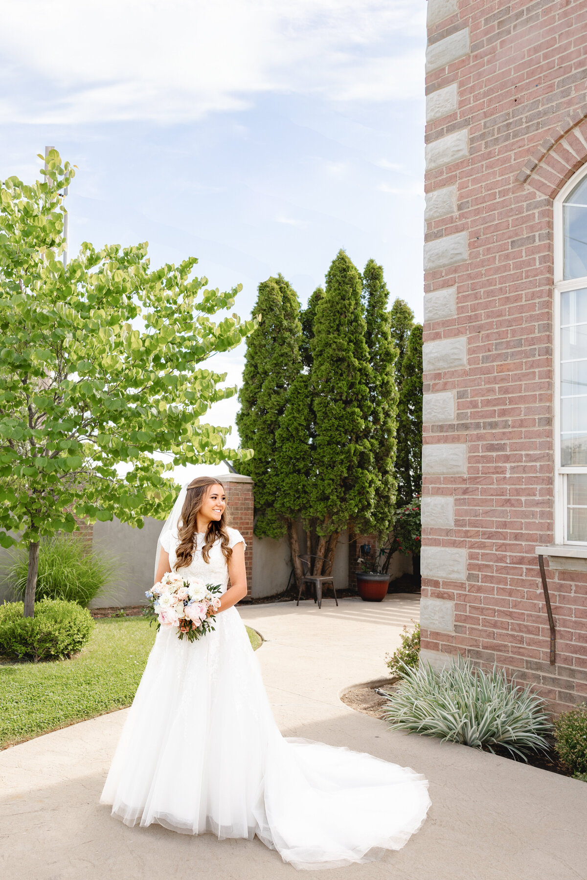 A bride in a flowing white gown holds a bouquet of soft pastel flowers, surrounded by vibrant greenery and an elegant brick venue.