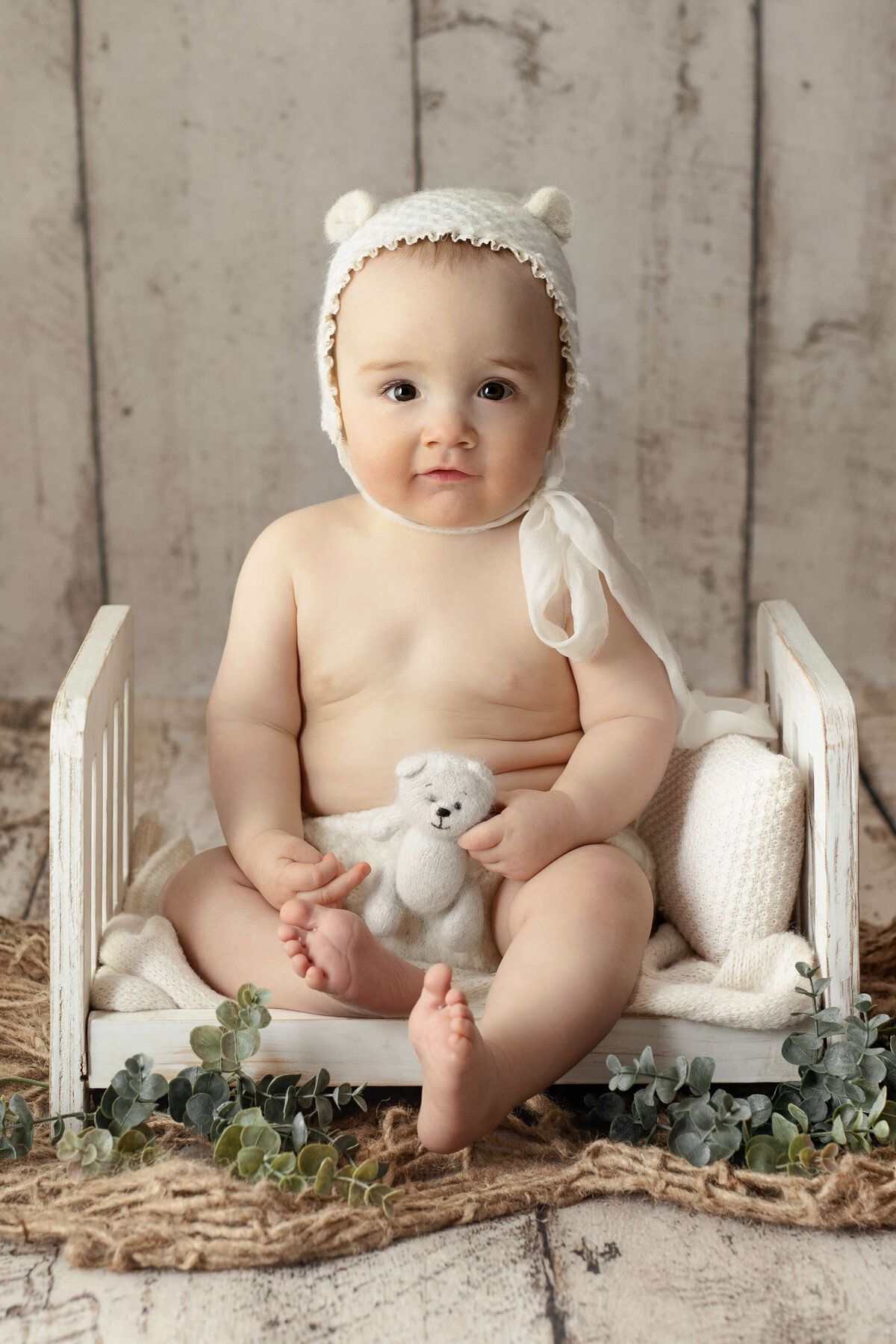 5 month old toddler wearing a bear bonnet sitting on a white wooden bed holding a white bear in front of a white wooden backdrop
