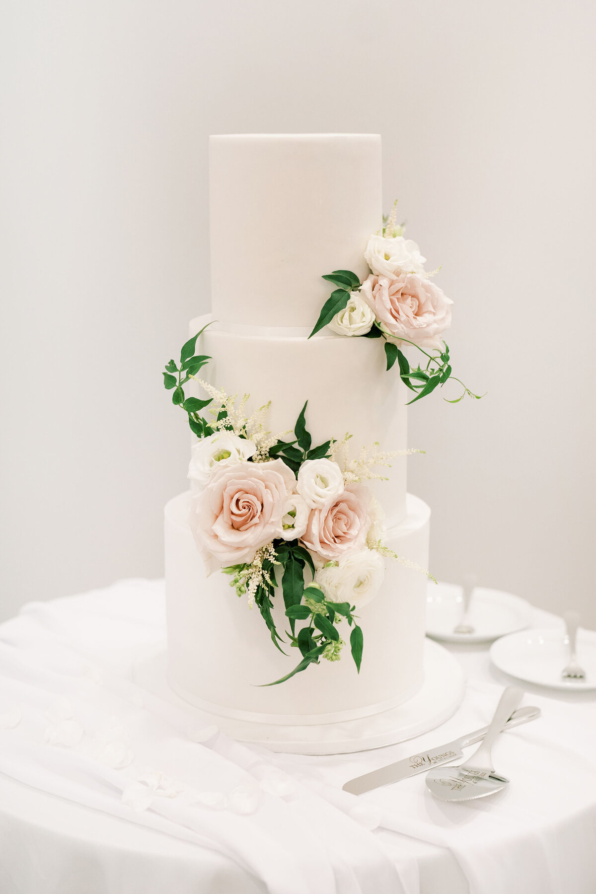 A three-tier white wedding cake adorned with pink and white roses and green foliage graces a table covered with a white cloth at this stunning Banff Alberta wedding. Cake-cutting utensils are nearby, ready for the couple to make their first slice.