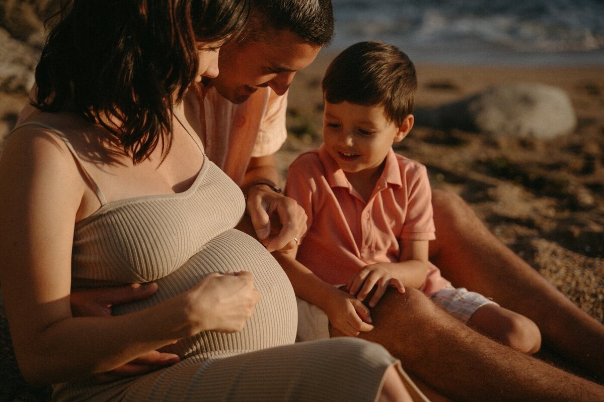 La famille assise sur le sable, le père et le fils touchent le ventre rond de la mère. Lumière dorée. Photo par Kamescop, photographe grossesse.