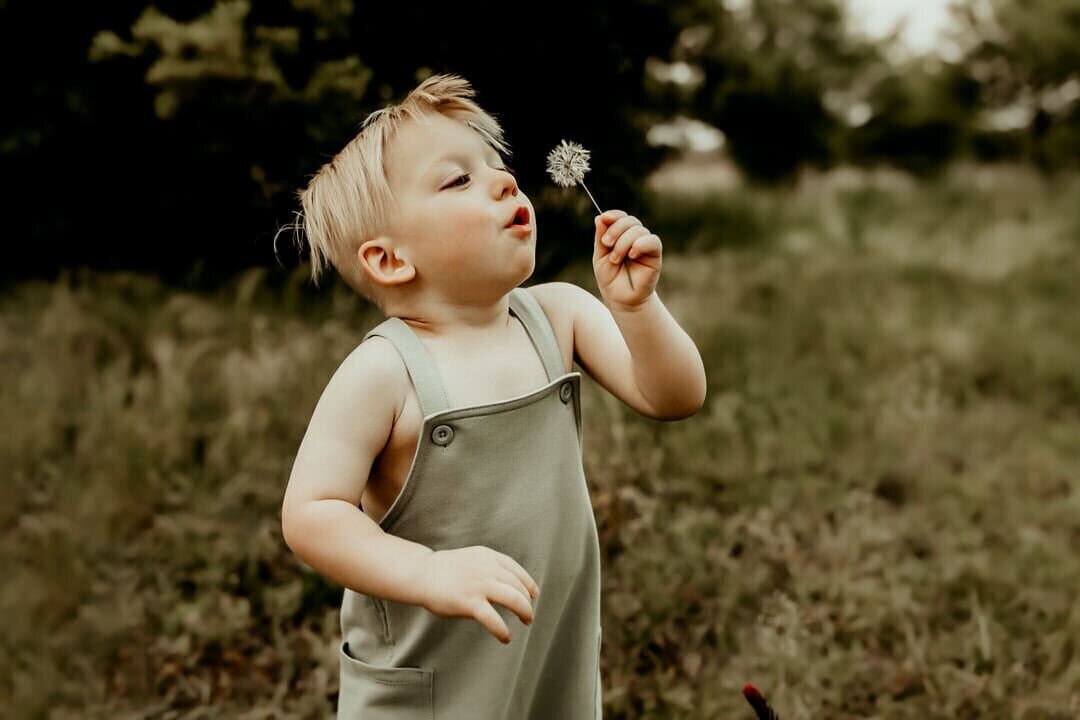A toddler blowing on a dandelion in a grassy field, wearing a green romper.