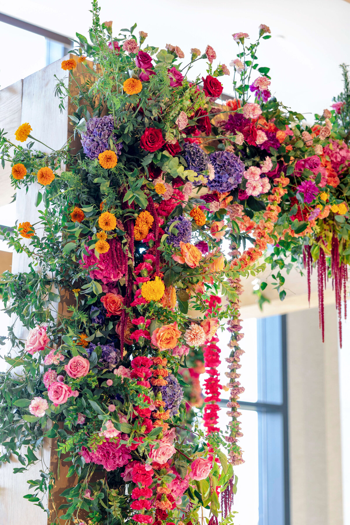 A vibrant floral arrangement with a variety of colorful flowers including roses, hydrangeas, and marigolds. The display is lush and dense, cascading down a wooden structure, set against a softly lit indoor background.